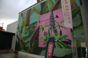 Street art de Mazza représentant une fresque végétale tropicale avec grandes feuilles vertes et fleur violette sur mur urbain.